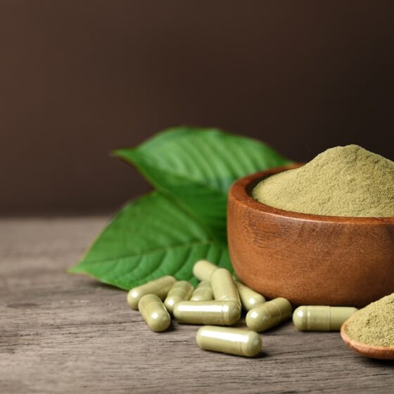 Kratom (Mitragyna speciosa) capsules with powder and fresh leaf on wood background.