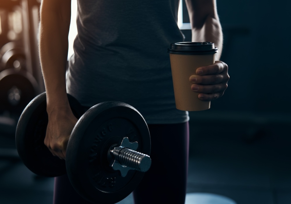 A fitness enthusiast holding a pre-workout coffee in one hand and a dumbbell in the other in gym for workout motivation.